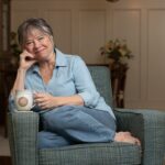 Mature woman sitting with her knees curled up on a blue chair enjoying a cup of tea
